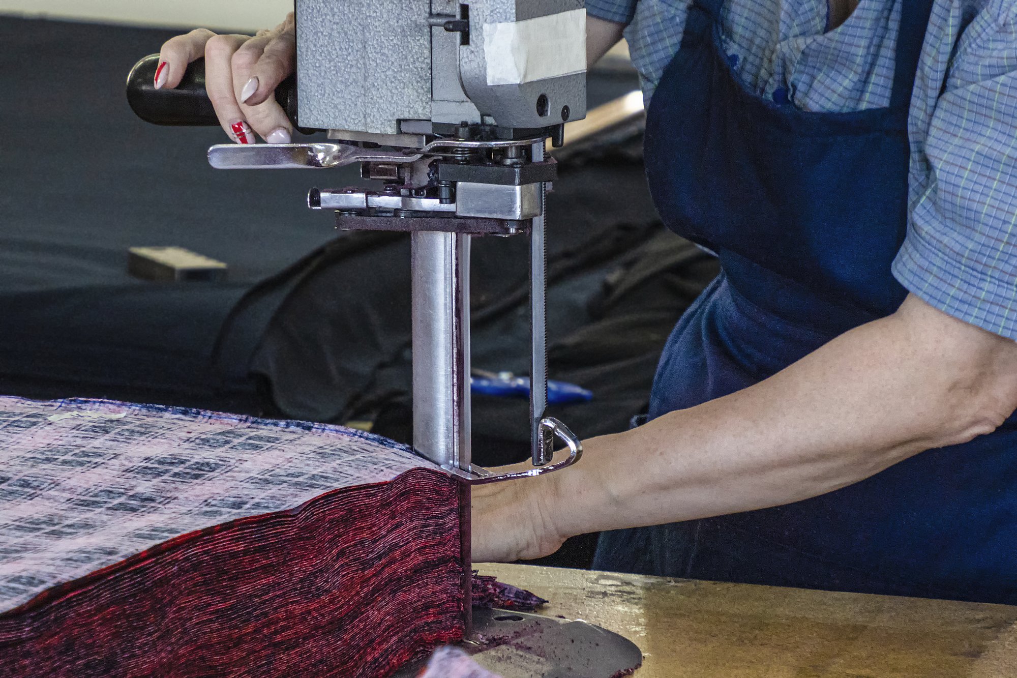 Fabricantes de ropa: primer plano de una persona que utiliza una máquina eléctrica para cortar tela sobre una mesa de trabajo, con un delantal azul y una camisa azul de cuadros y manga corta.