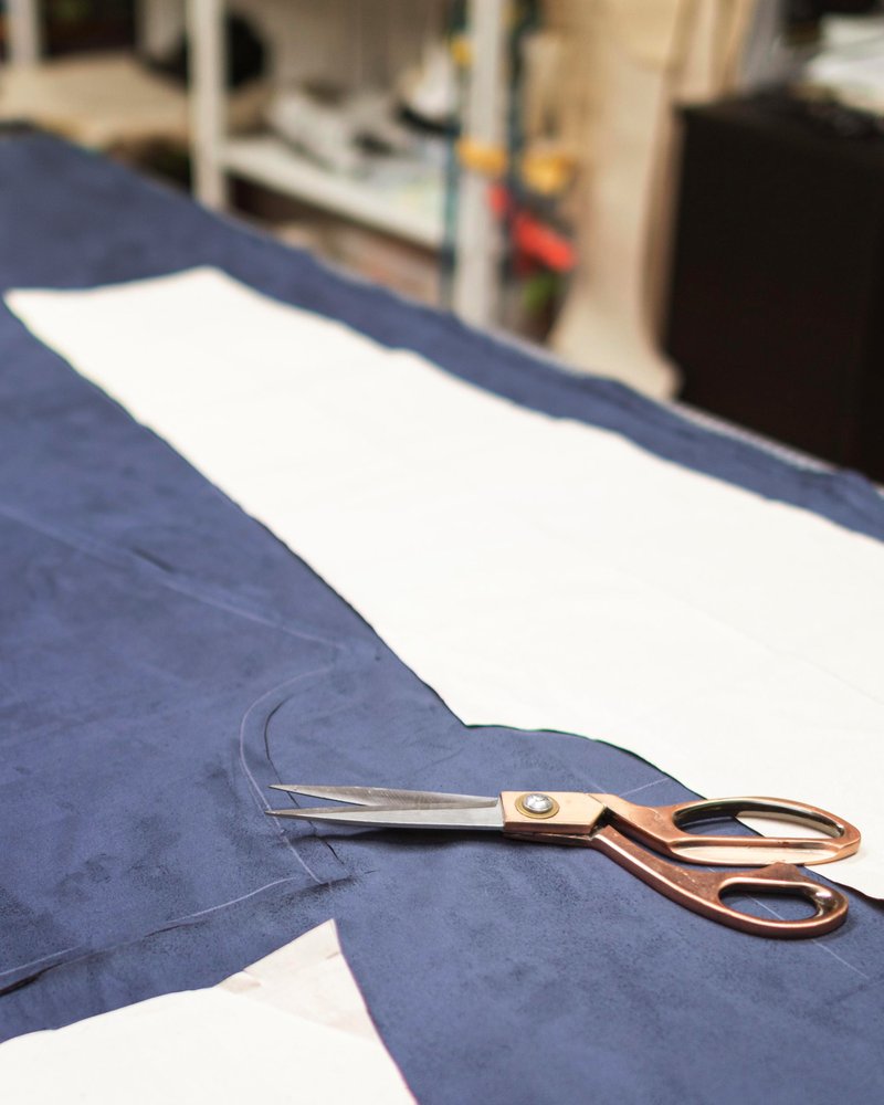 Un par de tijeras descansan sobre una tela azul oscuro con patrones de papel blanco clavados en ella, listas para cortar sobre una mesa de trabajo en un taller de costura o sastrería. En el fondo borroso se ven estanterías y diversos materiales.