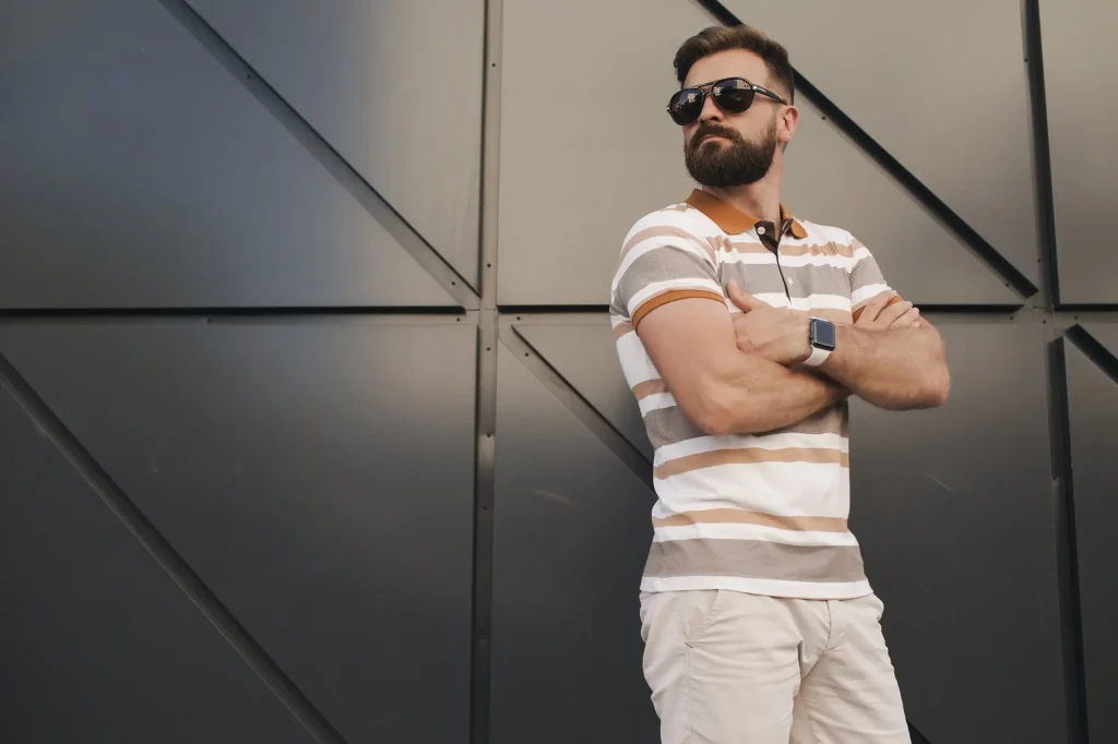 Un hombre con barba, con gafas de sol y camisa de rayas, está de pie con los brazos cruzados frente a una pared oscura y moderna. Lleva unos auriculares colgados del cuello y mira hacia un lado: vibraciones de estilo cerca de Inmobiliaria Grupo Sur en Fuengirola.