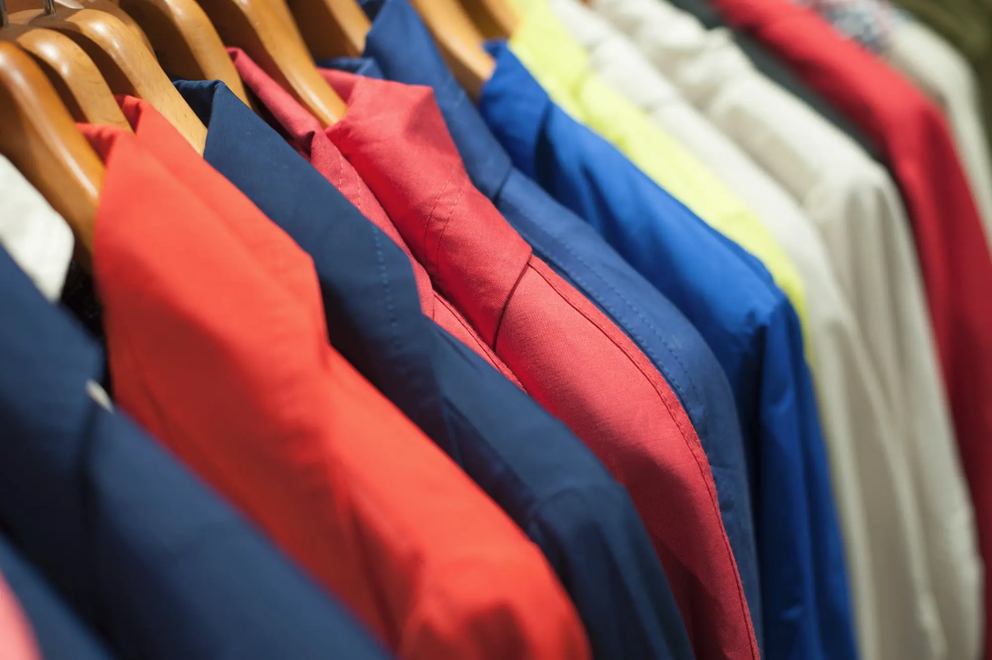 Una hilera de camisas de colores, entre ellas rojas, azules y blancas, cuelga ordenadamente de perchas de madera en un armario o en una tienda de ropa: perfecto para quienes buscan proveedores de ropa.
