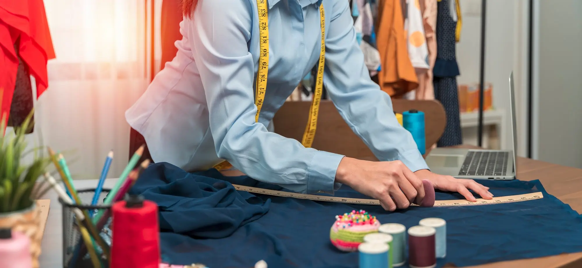 Una persona con camisa azul claro y una cinta métrica amarilla alrededor del cuello mide tela oscura sobre una mesa con herramientas de costura, carretes de hilo y ropa de fondo.