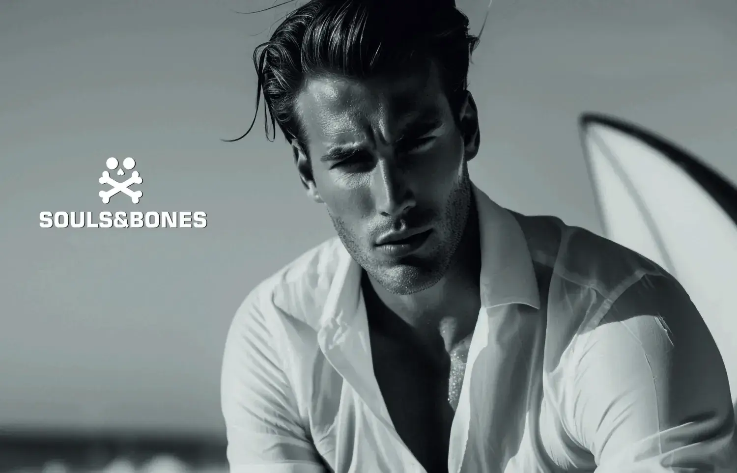 Un hombre con camisa blanca de cuello abierto mira intensamente a la cámara en una playa. La imagen es en blanco y negro, con el logotipo y el texto de Souls&Bones en el lado izquierdo. En el fondo se ve parcialmente una tabla de surf.